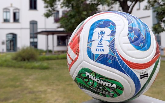 A close-up of a FIFA-branded soccer ball displaying colorful patterns and stars, positioned outdoors with a blurred background of a building and greenery.