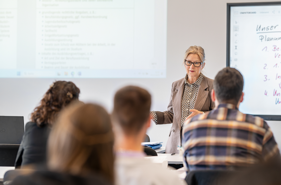 A teacher stands in front of a classroom, engaging with students seated at desks. A presentation is visible on a screen, with notes in German about planning and training processes. The focus is on the teacher's interaction with the class.
