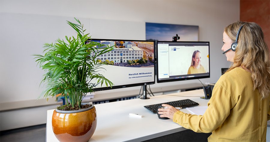 A woman with long hair and headphones sits at a desk, working on a computer with dual monitors. One screen displays a webpage with a building, while the other shows a video call with another woman. A potted plant adds a touch of greenery to the workspace.