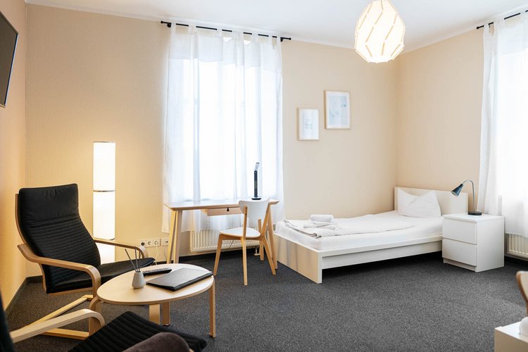 A cozy, modern bedroom featuring a single bed with white bedding, a small desk, and a comfortable chair. Soft natural light fills the space through large windows, complemented by light-colored walls and a dark carpet. A minimalist lamp and decorative wall art add subtle charm.