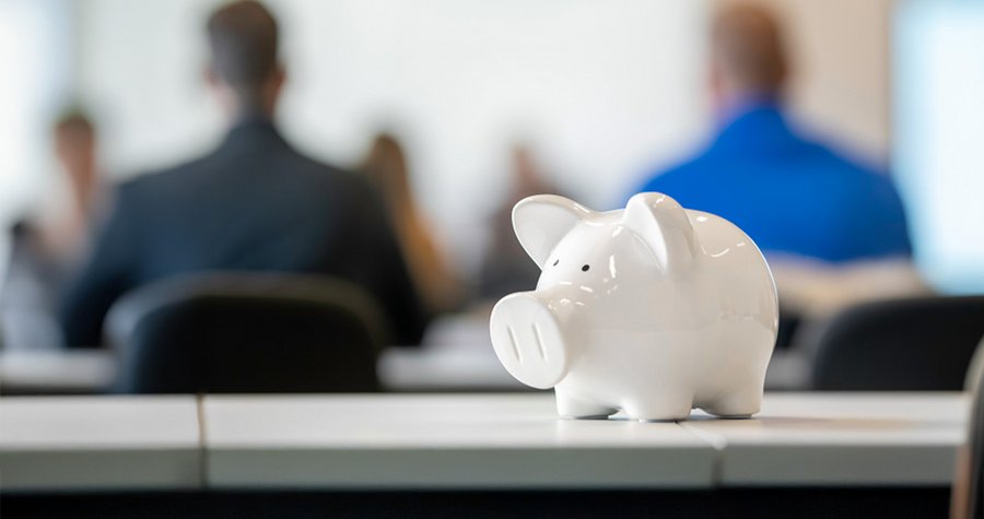 A white piggy bank is prominently displayed in the foreground, symbolizing saving and financial planning. In the background, a group of people is seated, out of focus, likely engaged in a discussion or presentation about finances.
