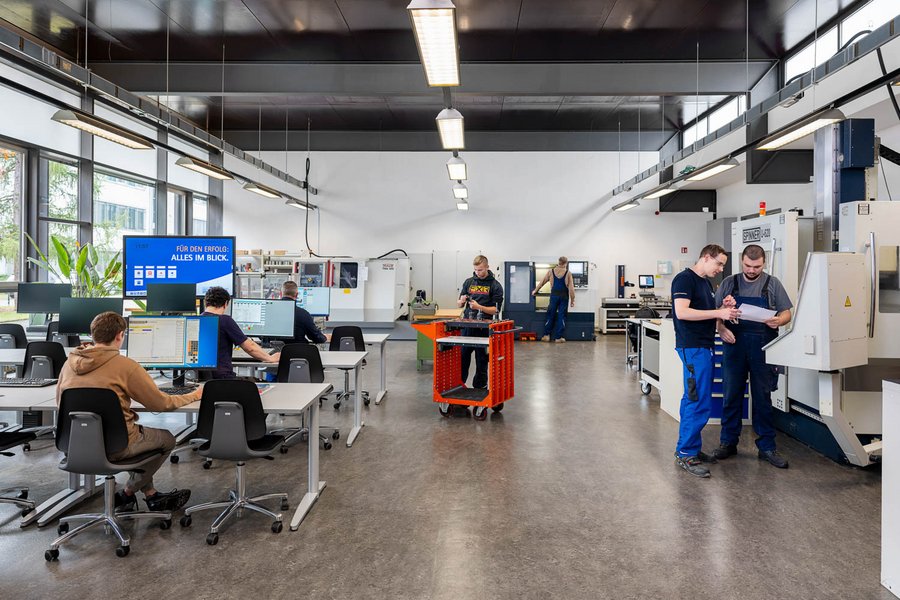 A modern workshop featuring several individuals engaged in various tasks. Some are working at computers, while others are collaborating and inspecting a project. Machinery and tools are visible throughout the space, emphasizing a productive environment focused on technical skill development and manufacturing.