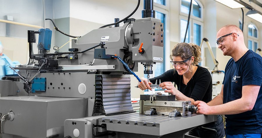 A woman and a man are working together at a milling machine in a workshop. Both are wearing safety goggles and focused on their task, with the woman operating the machine while the man assists her. The environment is well-lit with large windows in the background.