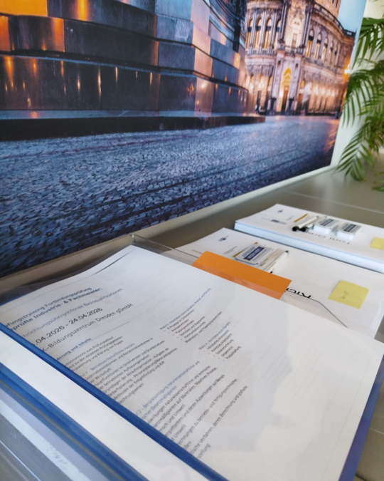 A clear view of an organized workspace featuring a binder with documents and papers, a pen, and a small notepad. In the background, there is a large photo of a building facade and cobblestone street. Green plant leaves are partially visible.