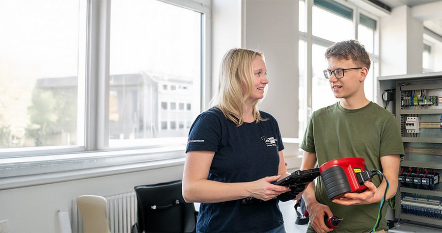 Two individuals, a woman and a man, are engaged in conversation in a bright, modern workspace. The woman holds a tablet, while the man is handling a device. A control panel with various electrical components is visible in the background.