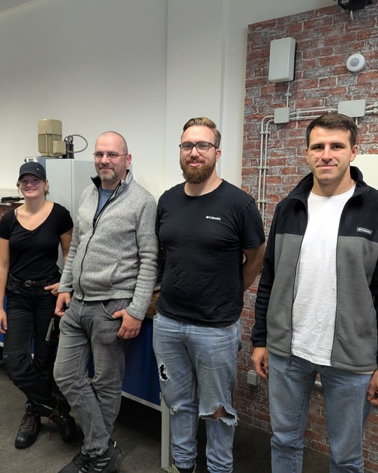 Four individuals stand side by side in a workshop setting. They are wearing casual work attire and appear engaged in a professional environment. The background features a brick wall and various equipment, suggesting a focus on teamwork or a collaborative project.