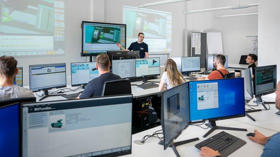 Ein dozierender Mann steht vor einer Gruppe von Teilnehmern in einem modernen Schulungsraum. Auf großen Bildschirmen werden technische Illustrationen und Software-Displays gezeigt. Die Atmosphäre ist konzentriert, während die Teilnehmer an ihren Computern arbeiten und dem Vortrag folgen.