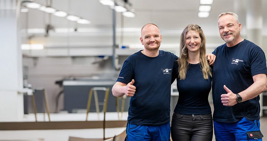 Three individuals are smiling and posing together in a well-lit industrial setting. They are dressed in matching navy blue shirts and casual attire, with two men giving thumbs up. The background features equipment typical of a workshop or manufacturing area.