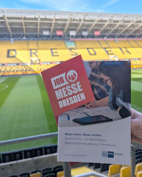 A person holds a pamphlet for a job fair in Dresden in front of an empty stadium. The pamphlet highlights opportunities for professional development and education, with text in both German and English. The stadium’s stands are visible in the background, emphasizing the event's location.