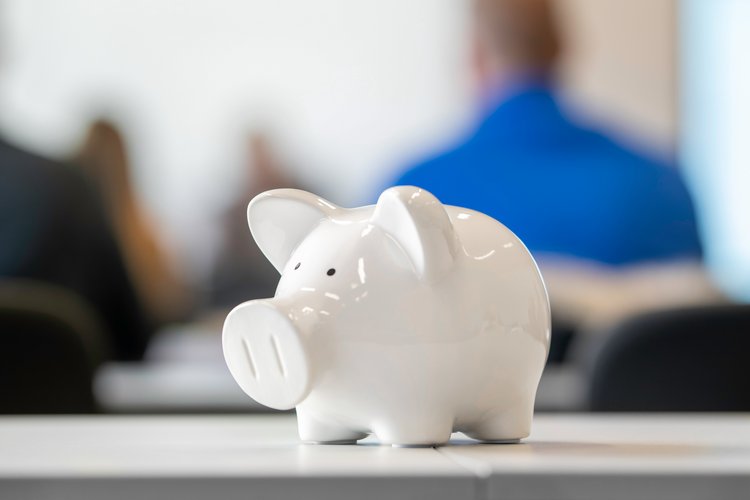A white piggy bank sits on a table in the foreground, symbolizing savings and financial planning. In the blurred background, a group of people can be seen attending a presentation or meeting.