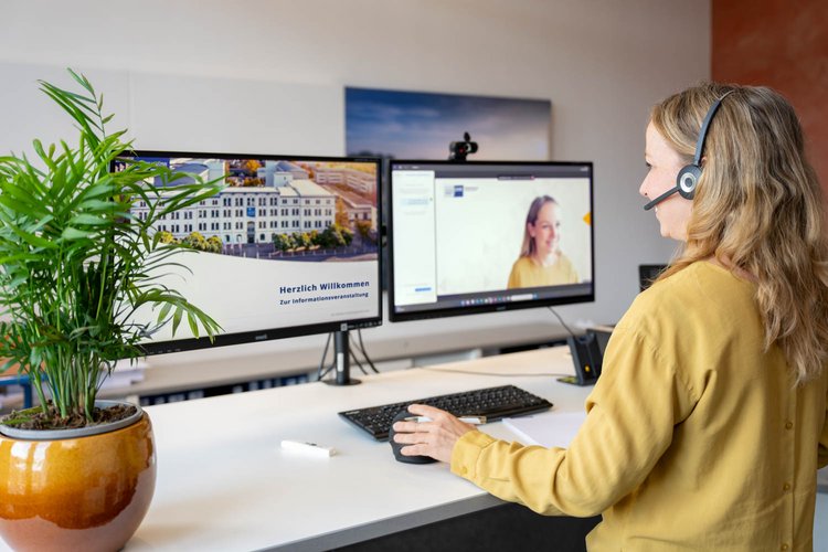 A woman with long hair is seated at a desk wearing a headset, engaging in a video call. Two computer monitors display a welcome message and the video call interface. A potted plant adds a touch of greenery to the workspace.