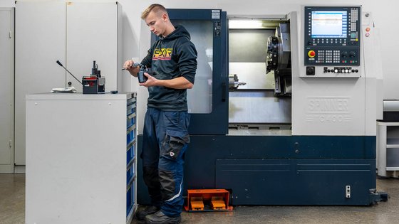 A technician stands in a workshop, holding a tool and inspecting it closely. Nearby, a computer-controlled milling machine is visible. The workspace includes a multi-drawer cabinet and various equipment, highlighting a focus on precision and machinery operations.