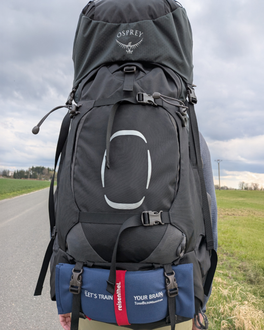 A person is seen from behind, wearing a large black backpack with the "Osprey" logo. The backpack has multiple straps and compartments, and a blue patch on the bottom reads "LET'S TRAIN YOUR BRAIN." The background features an empty road and green fields under a cloudy sky.