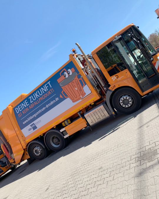 A side view of a bright orange garbage truck parked on a street. The truck features a large advertisement on its side, emphasizing sustainability with the text "Deine Zukunft ist kein Abfallprodukt" and a cartoonish illustration related to recycling.