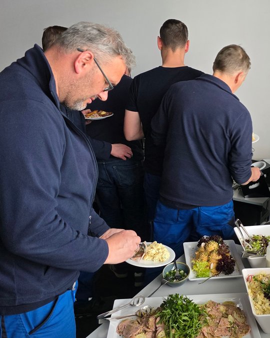 A group of people is serving food from a buffet. In the foreground, a man with glasses and gray hair is plating food on a white dish. He is focused on his meal, while others in dark clothing are filling their plates in the background.