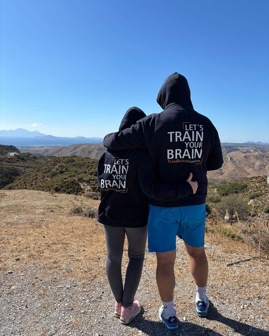 A couple stands together, overlooking a scenic landscape with mountains and a clear sky. They wear matching hoodies featuring the phrase “LET’S TRAIN YOUR BRAIN” on the back. The woman has her arm around the man's waist, creating a sense of connection and teamwork.