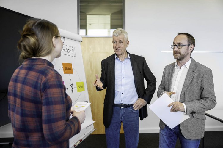 Three professionals are engaged in a discussion in an office setting. One person, wearing glasses and a blazer, holds a document while another, in a blue shirt, gestures as they speak to a woman in a plaid shirt. A flip chart displays various notes.